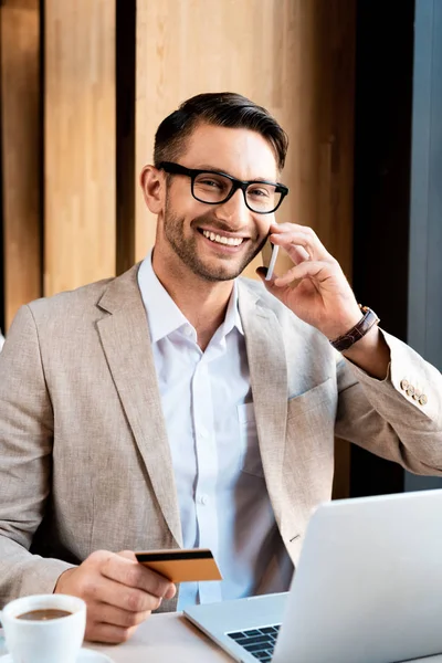 Smiling businessman in glasses talking on smartphone and holding credit card at table with laptop in cafe — Stock Photo