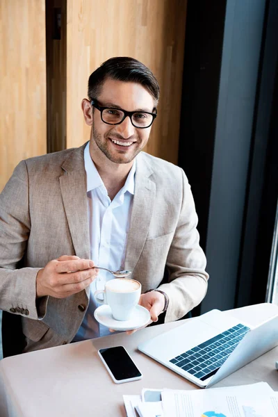 Улыбающийся бизнесмен в очках, держа чашку кофе, сидя за столом с ноутбуком в кафе — стоковое фото