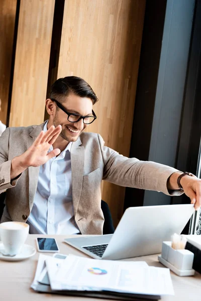 Excited businessman in glasses waving hand to laptop screen in cafe — Stock Photo