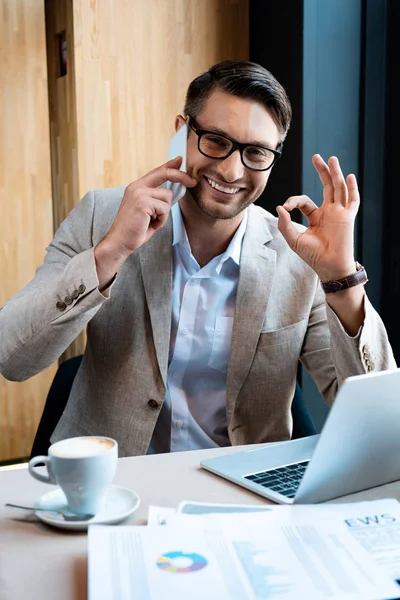 Uomo d'affari sorridente in occhiali parlando su smartphone e mostrando segno giusto in caffè — Foto stock