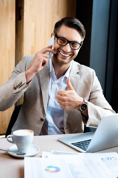Hombre de negocios sonriente en gafas hablando en smartphone y mostrando el pulgar hacia arriba en la cafetería - foto de stock