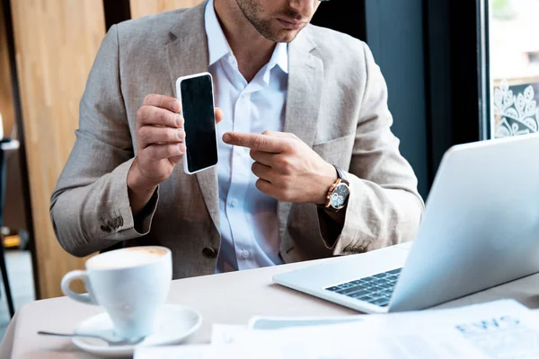 Обрезанный вид бизнесмена, указывающего пальцем на смартфон с пустым экраном перед ноутбуком в кафе — стоковое фото