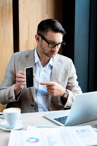 Homem de negócios apontando com o dedo para o smartphone com tela em branco na frente do laptop no café — Fotografia de Stock