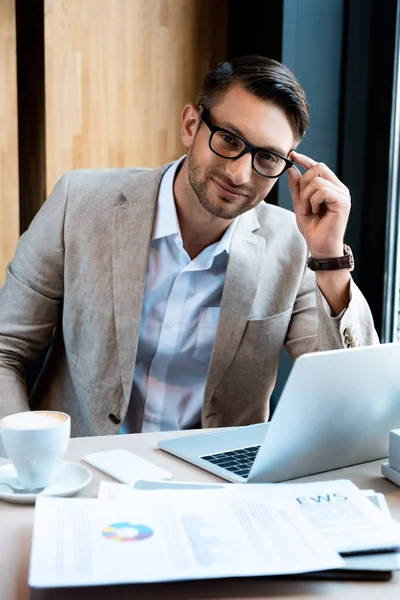 Smiling businessman in glasses sitting at table with laptop, documents and cup of coffee in cafe — Stock Photo