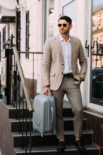 Vue pleine longueur de l'homme d'affaires en lunettes de soleil tenant valise sur la rue — Photo de stock