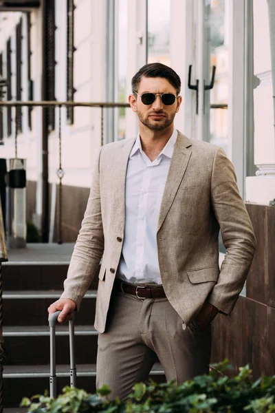 Hombre de negocios serio en gafas de sol sosteniendo la maleta y de pie con la mano en el bolsillo en la calle - foto de stock