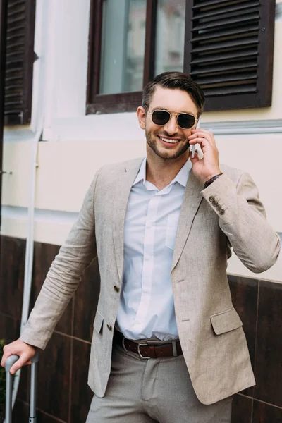 Улыбающийся бизнесмен в солнечных очках, держащий чемодан и разговаривающий по смартфону на улице — стоковое фото