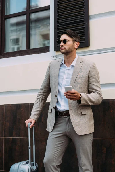 Businessman in sunglasses holding suitcase and smartphone on street — Stock Photo