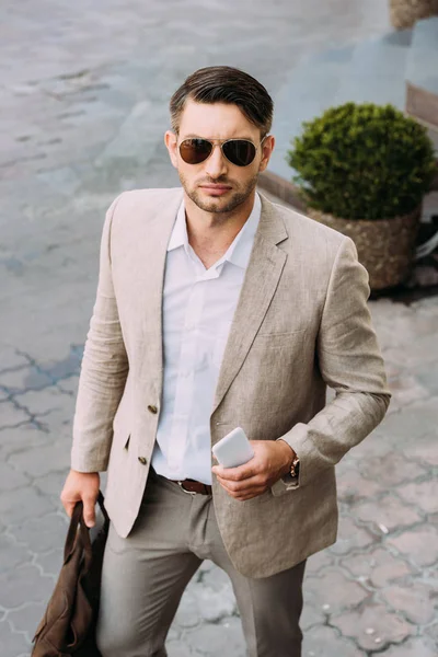 Serious businessman in sunglasses holding bag and smartphone on street — Stock Photo