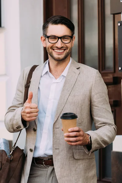 Vorderansicht eines lächelnden Geschäftsmannes mit einer Brille, die einen Coffee to go hält und den Daumen auf der Straße zeigt — Stockfoto