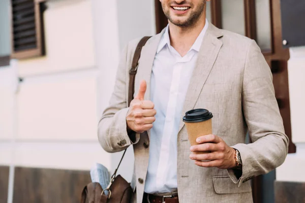 Recortado vista de sonriente hombre de negocios en vasos sosteniendo café para ir y mostrando el pulgar hacia arriba en la calle - foto de stock