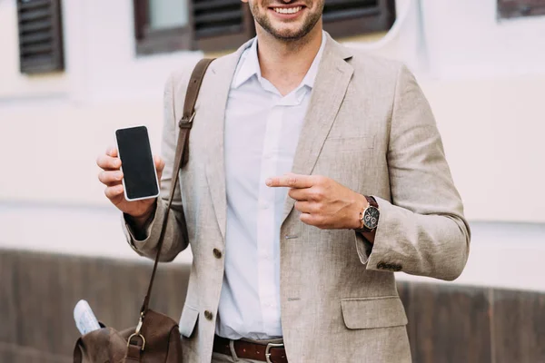 Recortado vista de sonriente hombre de negocios señalando con el dedo en el teléfono inteligente con pantalla en blanco en la calle - foto de stock