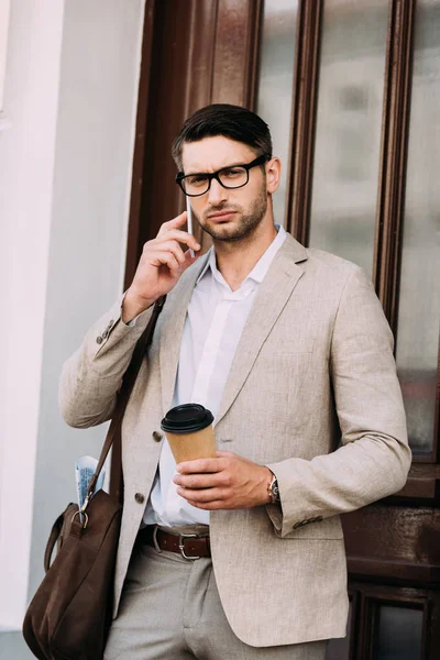 Hombre de negocios pensativo con bolsa de cuero hablando en el teléfono inteligente y sosteniendo el café para salir a la calle - foto de stock