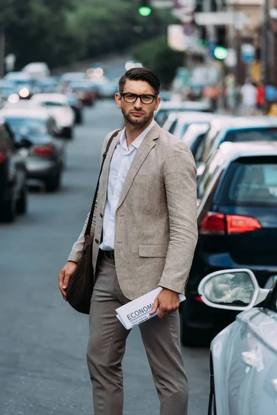 Homem sério em óculos segurando jornal na rua com carros — Fotografia de Stock