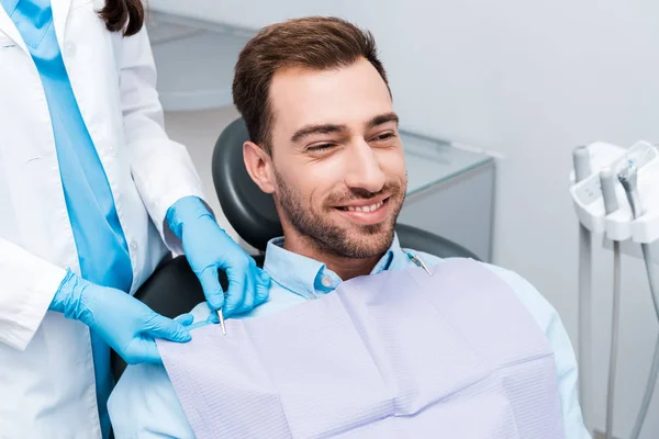 Vista recortada del dentista en guantes de látex azul de pie cerca de paciente alegre - foto de stock