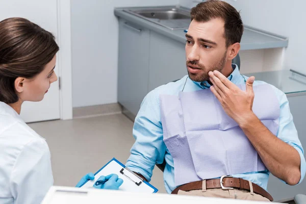 Selective focus of upset man gesturing and looking at attractive dentist in latex gloves holding pen and clipboard — Stock Photo