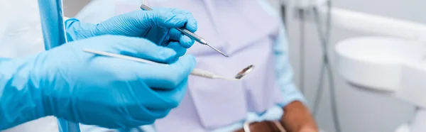 Prise de vue panoramique du dentiste en gants de latex bleu tenant des instruments dentaires — Photo de stock