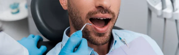 Tiro panorâmico do dentista segurando instrumento dentário perto do homem — Fotografia de Stock