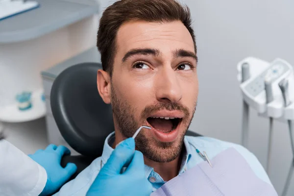 Vista cortada de dentista segurando instrumento dentário perto de homem bonito — Fotografia de Stock