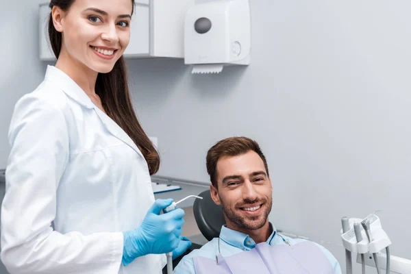 Привлекательный и счастливый стоматолог, держащий стоматологическое оборудование рядом с счастливым человеком — стоковое фото