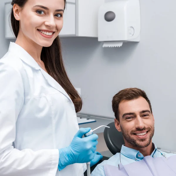 Beautiful and happy dentist holding dental equipment near happy man — Stock Photo