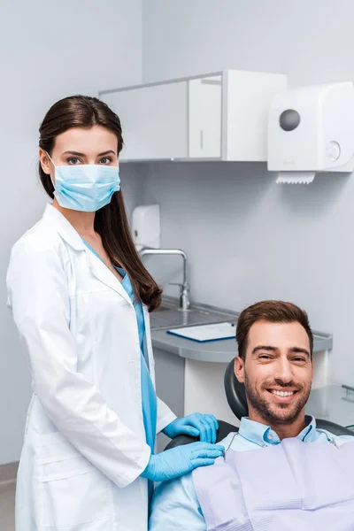 Стоматолог в медицинской маске и латексных перчатках, стоящих рядом с счастливым пациентом — стоковое фото