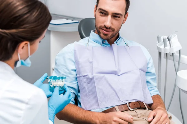 Foco seletivo de homem bonito olhando para dentista segurando modelo de dentes na clínica — Fotografia de Stock