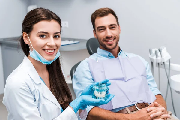 Селективный фокус привлекательного стоматолога в латексных перчатках, держащего модель зубов рядом с пациентом с сжатыми руками — стоковое фото