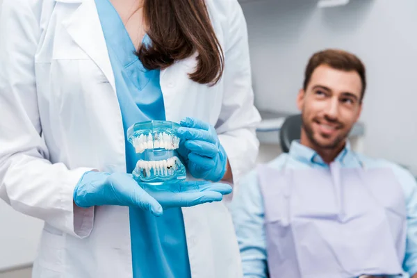 Corte vista de dentista segurando dentes modelo e de pé perto de homem feliz — Fotografia de Stock