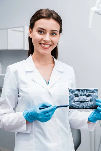 Alegre dentista sosteniendo pluma cerca de rayos X y sonriendo en la clínica - foto de stock