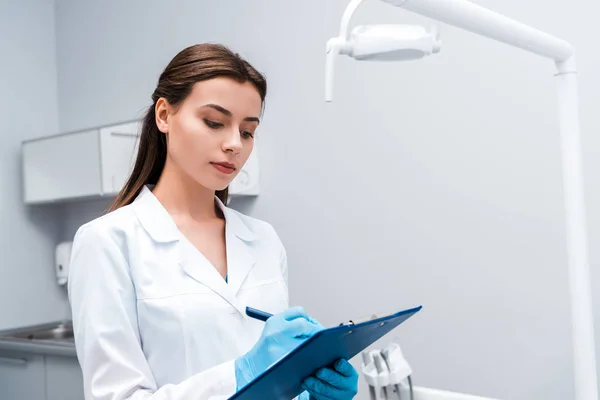 Atraente dentista escrever enquanto segurando prancheta na clínica — Fotografia de Stock