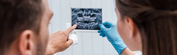 Plano panorámico del dentista sosteniendo rayos X cerca del paciente señalando con el dedo en la clínica - foto de stock