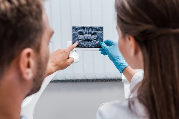 Обрезанный вид стоматолога, держащего рентген рядом с пациентом, указывающим пальцем в клинике — стоковое фото