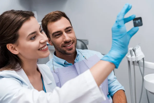 Селективное внимание привлекательного стоматолога и улыбающегося пациента, смотрящего на рентген — стоковое фото