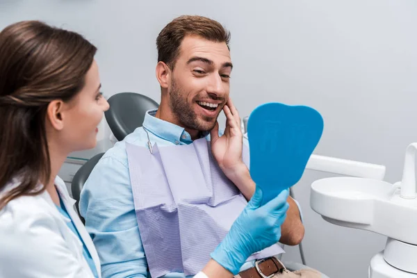 Selective focus of bearded patient touching face while holding mirror near dentist — Stock Photo