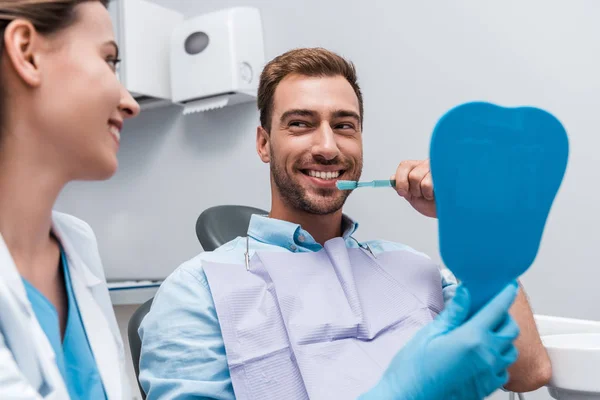 Enfoque selectivo de hombre feliz cepillarse los dientes cerca de atractivo dentista celebración espejo - foto de stock