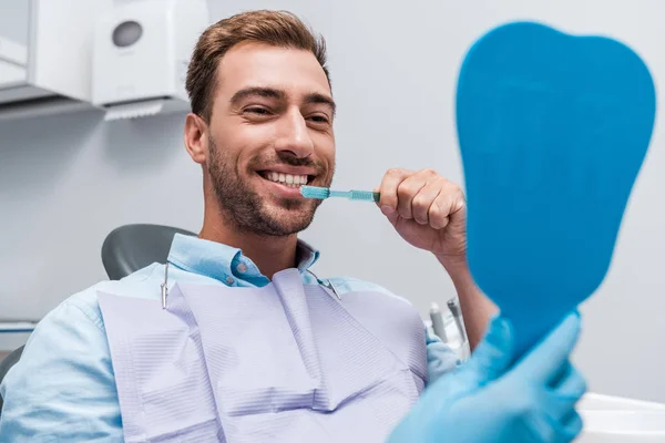 Cropped view of dentist holding mirror near cheerful man brushing teeth — Stock Photo