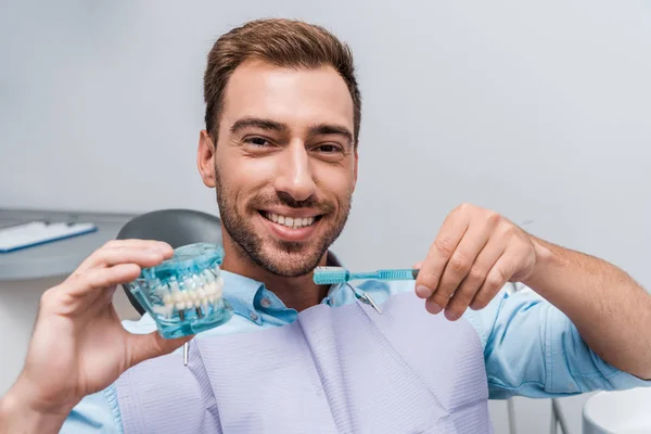 Hombre feliz sosteniendo modelo de diente y cepillo de dientes en la clínica - foto de stock