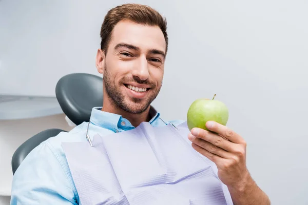 Веселий бородатий чоловік тримає зелене яблуко і посміхається в стоматологічній клініці — стокове фото