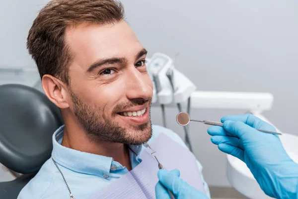 Vista recortada del dentista en guantes de látex que sostienen instrumentos dentales cerca del paciente - foto de stock