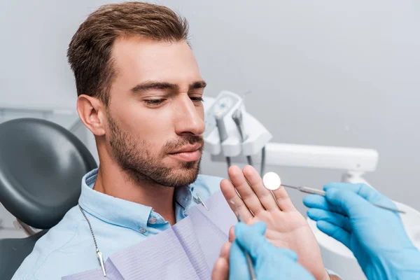 Vista recortada del dentista en guantes de látex que sostiene instrumentos dentales cerca del gesto del paciente en la clínica - foto de stock