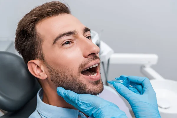 Vue recadrée du dentiste en gants de latex bleu tenant le dispositif de retenue près de l'homme barbu — Photo de stock