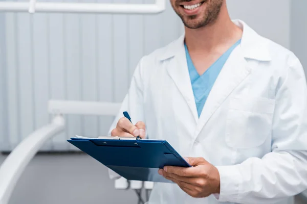 Обрезанный вид счастливого бородатого врача, держащего планшет и ручку в клинике — стоковое фото