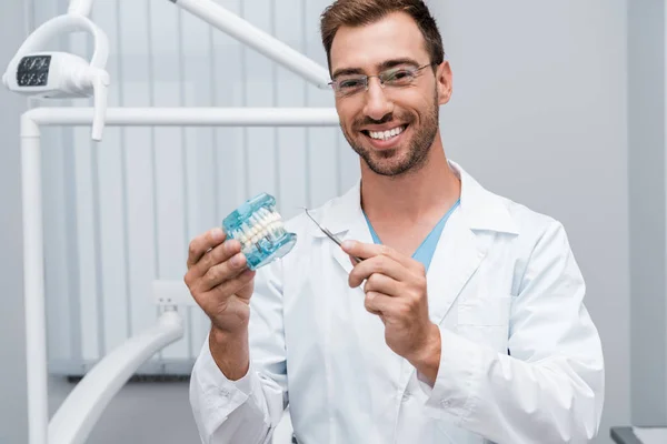 Dentista alegre em óculos segurando instrumento dental e modelo de dente nas mãos — Fotografia de Stock