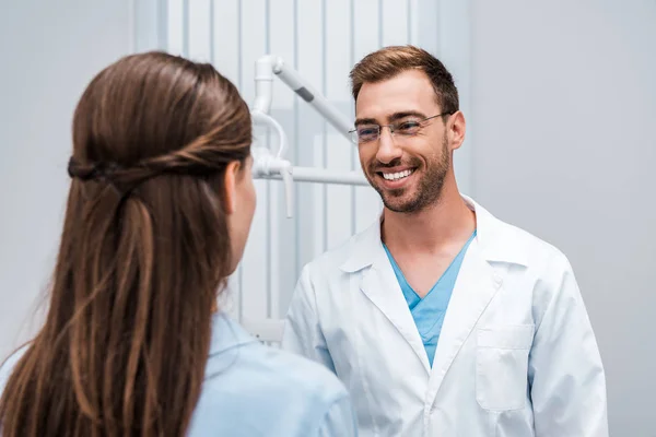 Messa a fuoco selettiva di dentista allegro in occhiali che guardano il paziente in clinica — Foto stock