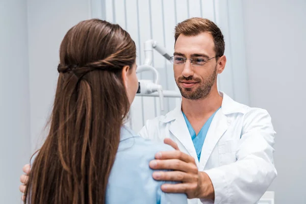 Селективный фокус жизнерадостного стоматолога в очках трогательных плеч пациента в клинике — стоковое фото