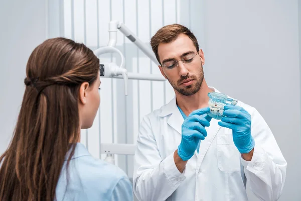 Enfoque selectivo del dentista barbudo en gafas que sostienen el modelo dental cerca del paciente - foto de stock