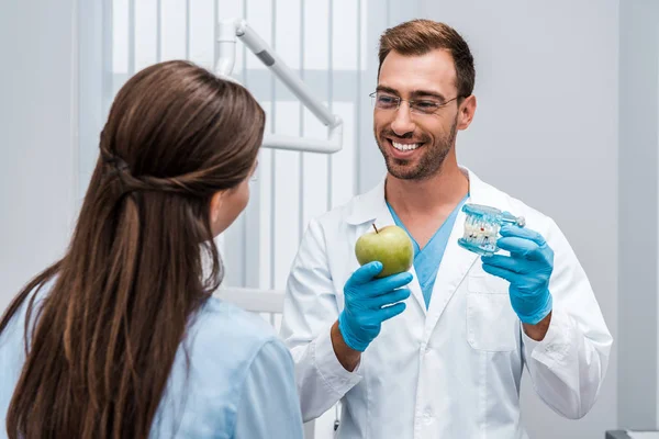 Enfoque selectivo del dentista feliz en gafas que sostienen el modelo de diente cerca del paciente - foto de stock