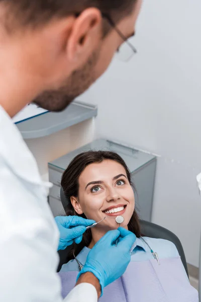 Messa a fuoco selettiva dell'uomo in guanti di lattice in possesso di strumenti dentali vicino alla donna attraente — Stock Photo