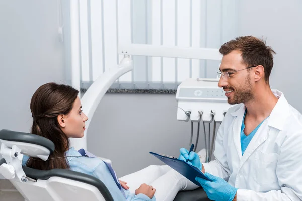 Handsome dentist in glasses holding pen and clipboard near attractive patient — Stock Photo